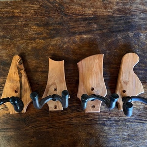 May include: Four wooden guitar wall hangers with black metal supports. The wooden hangers vary in shape and wood grain, with two featuring a lighter wood and two with a darker, more textured appearance. Each hanger has two curved supports.