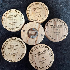 Puede incluir: Conjunto de cinco abridores de botellas de madera con un diseño de U.S. Geological Survey. Cada abridor presenta un parque nacional diferente: Arches, Capitol Reef, Bryce Canyon, Zion y Canyonlands. Los abridores están grabados con el nombre del parque, la fecha de "Establecimiento" y "Bench Mark".