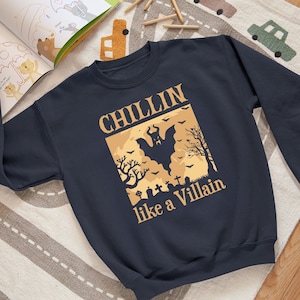 May include: A navy blue sweatshirt with a graphic print of a bat flying over a graveyard. The text "Chillin' like a Villain" is printed below the graphic.
