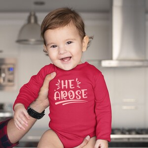 May include: A red long-sleeve baby onesie with the text "HE' AROSE" in pink lettering.