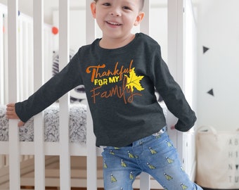 May include: A young child wearing a dark grey long-sleeved shirt with the text "Thankful for my family" and a yellow leaf graphic. The child is standing in front of a white crib and wearing blue jeans with a yellow banana pattern.