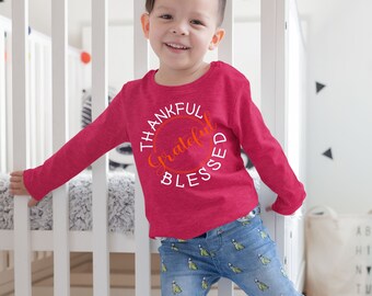 May include: A young child wearing a pink long-sleeved shirt with the text "Thankful Grateful Blessed" in a circle. The child is standing in front of a white crib.