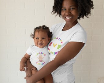 Puede incluir: Una madre y su hija llevan camisetas blancas a juego con el texto "Amazing IVF Warrior" y "Amazing IVF Victory" en colores arcoíris. La hija está siendo sostenida por la madre.