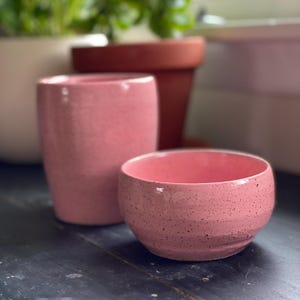 May include: A tall cylindrical pink ceramic cup and a matching shallow pink ceramic bowl, both featuring a speckled finish with dark brown flecks throughout the smooth, glossy glaze, arranged together on a dark surface.