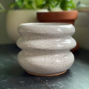 May include: A cylindrical ceramic planter with a glossy off-white glaze and dark brown speckles throughout. The vessel features three distinct horizontal rounded ridges encircling the body, creating a stacked, undulating profile.