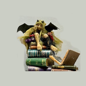 May include: A green and black fairy figurine with bat wings sits on a stack of vintage books. The fairy is holding a red book and has a mischievous expression.
