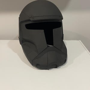 May include: A dark grey helmet with a visor opening and a geometric design. The helmet is made of a hard material and rests on a white surface. The helmet's design features a rounded top and a detailed front.