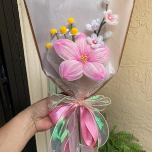 May include: A bouquet of handcrafted pipe cleaner flowers wrapped in clear plastic with a pink iridescent ribbon. The arrangement features a large pink central flower, yellow pom-pom blossoms, and small white flowers with pink centers.