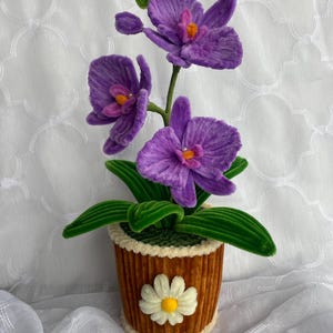 May include: A handcrafted purple orchid plant made from fuzzy pipe cleaners, featuring three vibrant blooms with orange centers. The plant is set in a brown, ribbed, cylindrical pot decorated with a single white daisy and cream-colored trim.