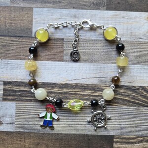 May include: A silver-tone chain bracelet featuring a variety of beads in yellow, black, brown, and cream colors. The bracelet includes two dangling charms: one shaped like a pirate figure with a red bandana and one shaped like a ship's wheel.