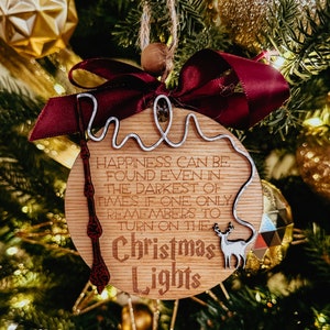 Puede incluir: Un adorno navideño de madera con una cinta roja y una inscripción de alambre plateado que dice "La felicidad se puede encontrar incluso en los momentos más oscuros si uno solo recuerda encender las luces de Navidad".
