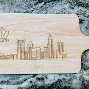 May include: A wooden cutting board with a laser-engraved design of the Charlotte skyline. The board has a handle and the text "CHARLOTTE" is engraved above the skyline.