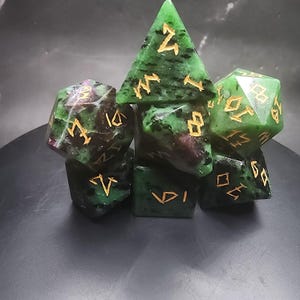 Puede incluir: Un juego de dados de piedras preciosas en varias formas, incluyendo una pirámide, un cubo y un d20. Los dados son verdes con inclusiones rojo oscuro y marcas doradas. El conjunto se muestra sobre una superficie oscura.