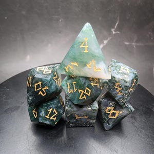 Puede incluir: Un conjunto de siete dados poliédricos para juegos de mesa. Los dados son una mezcla de verde y blanco con números dorados. Los dados están hechos de un material translúcido, y los números están en una fuente estilizada.