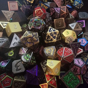 May include: A collection of polyhedral dice in various colors and designs. The dice include D4, D6, D8, D10, D12, and D20 shapes, with numbers visible on the faces. Colors range from gold and silver to red, purple, and green. Some dice have intricate patterns.