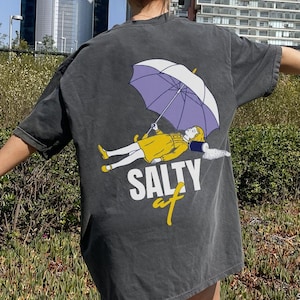 May include: Dark grey oversized t-shirt with a graphic on the back showing a person under a purple umbrella and the word "SALTY" in white and yellow. The shirt is worn by a person outdoors.