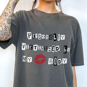 May include: A dark gray t-shirt with the text "PERSONALIY VICTIMIZED BY MY BIDY" printed on it in black and white letters. A red lip print is between the words "MY" and "BIDY".