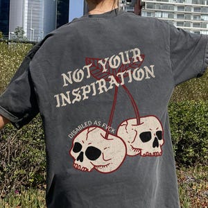 May include: Dark gray oversized t-shirt with the text "NOT YOUR INSPIRATION" above a graphic of two skull cherries. The words "DISABLED AS FVCK" are below the cherries. The shirt is worn outdoors.