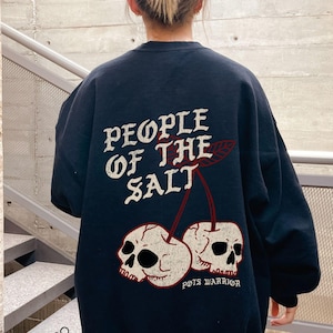 May include: Black sweatshirt with white text reading "PEOPLE OF THE SALT" and a graphic of two skulls as cherries. The text "POTS WARRIOR" is below the cherries. The sweatshirt is worn by a person.