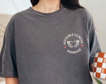 May include: Dark grey t-shirt with the text "INVISIBLE ILLNESS WARRIOR" and a heart-shaped graphic. The shirt is paired with blue jeans and a checkered orange and white mug. Decorative flowers are in the background.