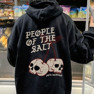 Peut inclure: Un sweat à capuche noir avec l'inscription "PEOPLE OF THE SALT" et un motif de deux crânes en forme de cerises. La personne se trouve devant un réfrigérateur de magasin.