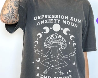 May include: A dark grey t-shirt with a white graphic of a frog meditating under a mushroom cap, surrounded by moons and stars. Text includes "DEPRESSION SUN ANXIETY MOON ADHD RISING". The shirt also has the text "ORIGINAL DESIGNER" and "hot girls don't buy knock-offs".