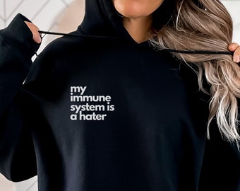 May include: Black hooded sweatshirt with the text "my immune system is a hater" in white. The sweatshirt has a front pocket and a drawstring hood. The image also includes text that says "S-5XL IN 6 COLORS" and "XL SIZE".