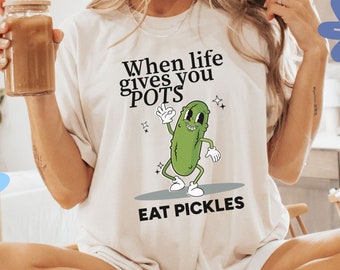 May include: A white t-shirt with the text "When life gives you pots eat pickles" and a cartoon pickle character. The pickle is green and has a white face with black eyes and a smile. The pickle is wearing white shoes and gloves. The t-shirt is being worn by a person sitting on a chair.