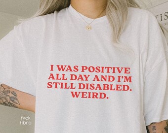 May include: White t-shirt with red text that reads "I was positive all day and I'm still disabled. Weird."