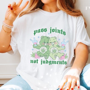 May include: White t-shirt featuring a graphic of two green cartoon bears holding beakers and cannabis leaves. The text "pass joints not judgments" is printed in green. The shirt is a casual style.