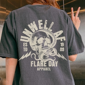 May include: Dark gray t-shirt with a distressed graphic on the back featuring a skull entwined with a snake, lightning bolts, and the text "UNWELL" and "FLARE DAY APPAREL". The shirt has a vintage, worn look.