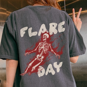 Peut inclure: T-shirt gris foncé avec un motif "FLARE DAY" vieilli. Le motif représente un squelette rouge avec des flammes. Le t-shirt a un aspect vintage et usé.