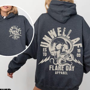 Könnte beinhalten: Dunkelgrauer Kapuzenpullover mit einem verwitterten Grafikdesign auf dem Rücken, das einen Totenkopf mit einer Schlange, den Text "UNWELL FLARE DAY APPAREL" und die Jahreszahlen "1989" zeigt. Auf der Vorderseite befindet sich eine kleinere Version des Designs.