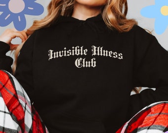 May include: Black hoodie with the text "Invisible Illness Club" in a gothic font. The person is wearing red and white plaid pajama pants. Decorative flowers are in the background. The Etsy shop URL is visible at the bottom.