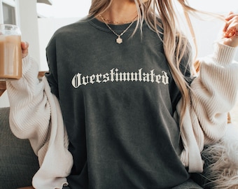 Puede incluir: Una mujer lleva una sudadera gris oscura con el texto "Overstimulated" en letras blancas. Está sentada en una silla de pelo blanco y sostiene un vaso de café helado.