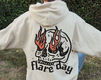 Puede incluir: Una sudadera con capucha de color crema con un gráfico de calavera y llamas. El texto "flare day" está impreso debajo de la calavera.