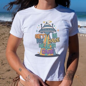 May include: White t-shirt with a graphic of a flying saucer and the text "Get in We Have Health Care".