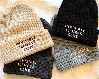 May include: Four knit beanies in beige, black, dark gray, and light gray, each with the text "INVISIBLE ILLNESS CLUB" embroidered in white. The hats are displayed on a fluffy white surface.