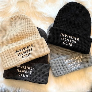 May include: Four knitted beanies in beige, black, dark grey, and light grey, each embroidered with the white text "INVISIBLE ILLNESS CLUB". The hats are arranged on a fluffy white surface.