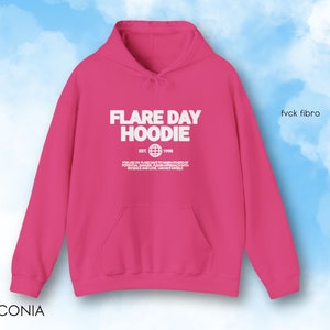 May include: A bright pink hoodie with the text "FLARE DAY HOODIE" in white. The hoodie has a front pocket and a drawstring hood. The text includes the year "EST. 1998" and a globe graphic. The text "fvck fibro" is on the right side.