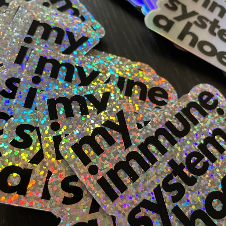 May include: A stack of silver holographic stickers with the text "i.my.immune.system.is.my" printed in black. The stickers are made of a shiny, iridescent material.