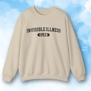 May include: A beige sweatshirt with the text "Invisible Illness Club" printed on the front in black.