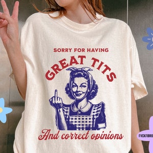 Puede incluir: Camiseta color crema con el texto "SORRY FOR HAVING GREAT TITS And correct opinions" en rojo y azul. El diseño presenta una ilustración vintage de una mujer haciendo un gesto obsceno. La camiseta tiene un corte holgado.