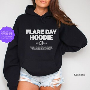 May include: Black hoodie with the text "FLARE DAY HOODIE" in white, along with "EST. 1998" and a globe graphic. The hoodie also has additional text below the main design. The image includes a purple sticker that says "ORIGINAL DESIGNER".