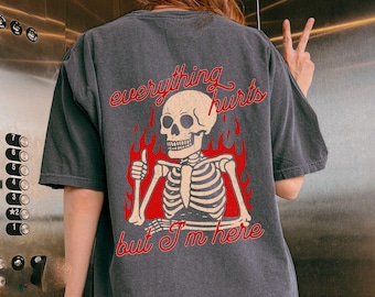 May include: Dark gray t-shirt featuring a skeleton graphic with red flames and the text "everything hurts but I'm here." The skeleton is giving a thumbs up. The shirt has short sleeves and a crew neck.