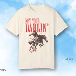 Puede incluir: Una camiseta de color crema con un gr&aacute;fico rojo de una persona montando un caballo. El gr&aacute;fico dice "Not Your Darlin'" en rojo.