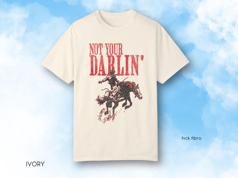 Puede incluir: Una camiseta de color crema con un gr&aacute;fico rojo de una persona montando un caballo. El gr&aacute;fico dice "Not Your Darlin'" en rojo.