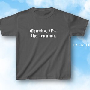Pu&ograve; includere: Una maglietta grigia scura con il testo "Thanks, it's the trauma." stampato in bianco sul davanti. Il testo &egrave; in font gotico. La maglietta ha un collo rotondo e maniche corte.
