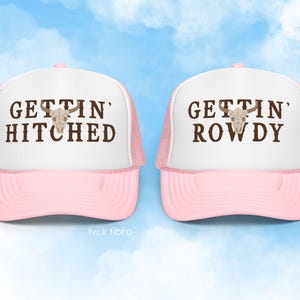 K&ouml;nnte beinhalten: Zwei rosafarbene Trucker-Caps mit wei&szlig;en Netzr&uuml;cken. Die linke Kappe tr&auml;gt die Aufschrift "Gettin' Hitched" und die rechte Kappe die Aufschrift "Gettin' Rowdy". Beide Kappen zeigen einen braunen Sch&auml;del mit H&ouml;rnern.