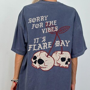 May include: A blue t-shirt with the text "SORRY FOR THE VIBES IT'S FLARE DAY" in white and red. The design features two skulls in the shape of cherries. The shirt is a casual, oversized fit.